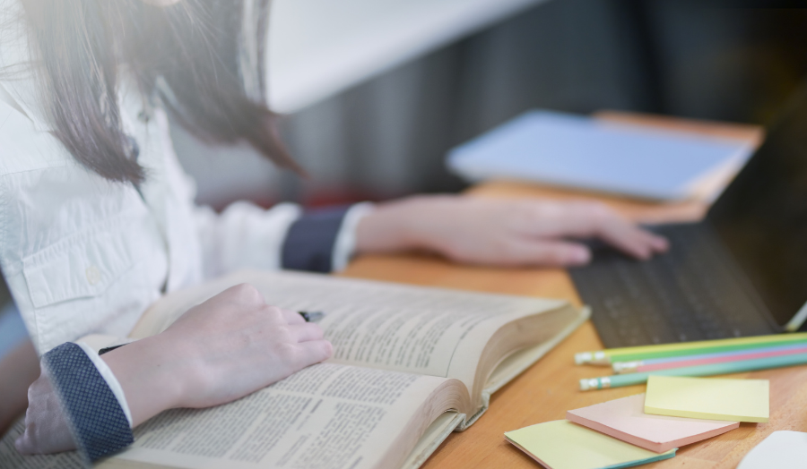 A student sits in front of an open book and a computer