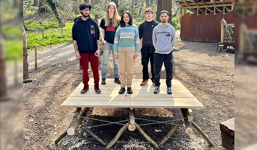 Five students stand on top of a wooden deck in a woodland clearing